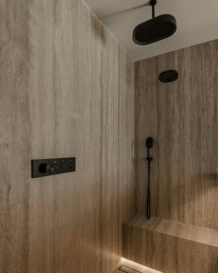 Photo d’illustration : une douche à l’italienne habillée de pierre claire, avec une banquette, une douche de tête et un mitigeur encastré noir.