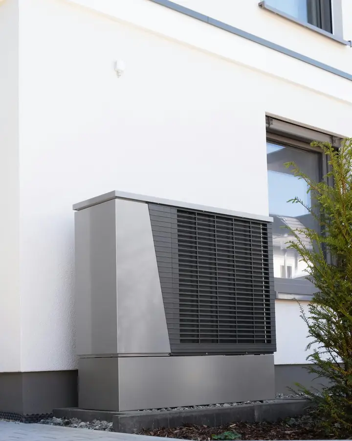 Photo d’illustration : l’unité extérieure d’une pompe à chaleur posée sur un socle, contre la façade d’une maison récente.