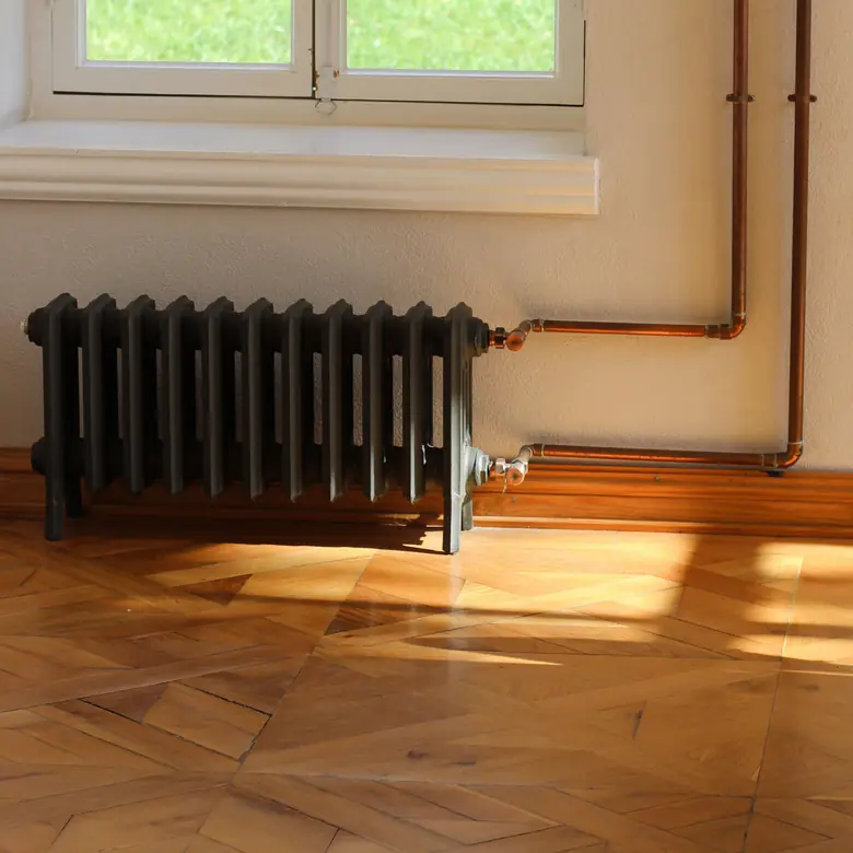 Photo d’illustration : un radiateur en fonte sous une fenêtre, raccordé par des tubes de cuivre apparents, dans une pièce au parquet baigné de soleil.