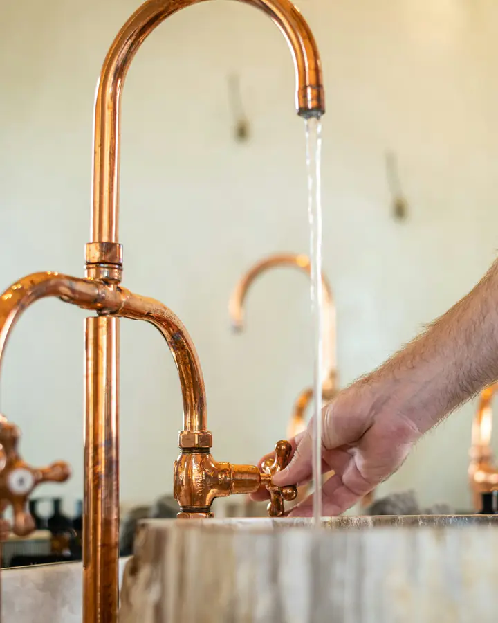 Photo d’illustration : de l’eau coule d’un robinet col de cygne en cuivre, qu’une main ouvre par sa poignée en croix, au-dessus d’une vasque en pierre.
